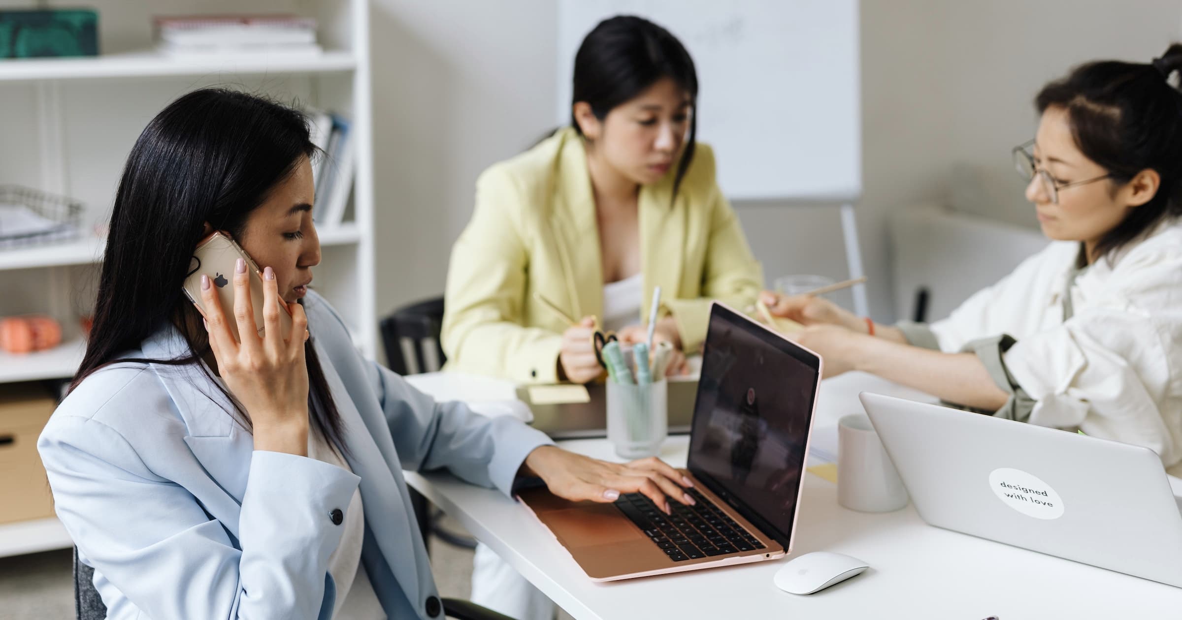 Three women in business attire working at a desk with laptops, one is on the phone